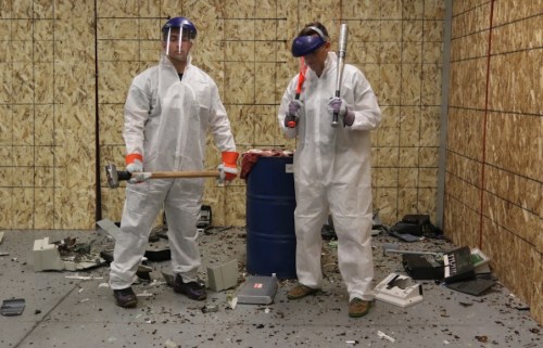 Two people in protective gear stand in a messy room, holding tools, ready to smash old electronics.