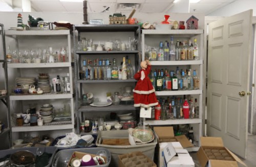 A cluttered room filled with shelves of glassware, bottles, and various items, with a Santa decoration hanging.