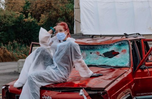A person in a protective suit and mask sits on a damaged red car with a shattered windshield.