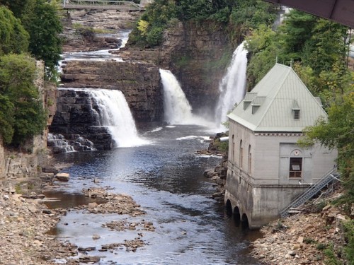 A scenic view of waterfalls cascading into a river beside a historic building surrounded by lush greenery.