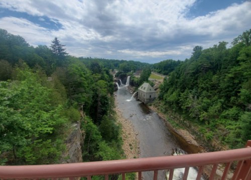 A scenic view of a river with waterfalls, surrounded by lush greenery and a historic building along the bank.