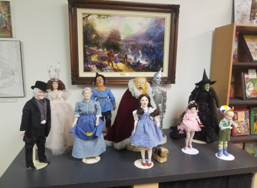 A collection of dolls representing characters from "The Wizard of Oz," displayed on a shelf with a scenic painting in the background.