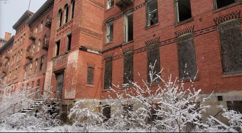 Abandoned brick building with boarded windows, surrounded by snow-covered shrubs and a winter landscape.