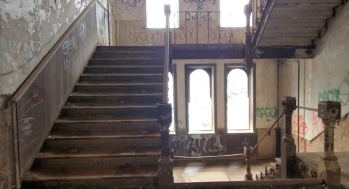 Abandoned staircase in a dilapidated building, with graffiti on walls and large windows letting in natural light.