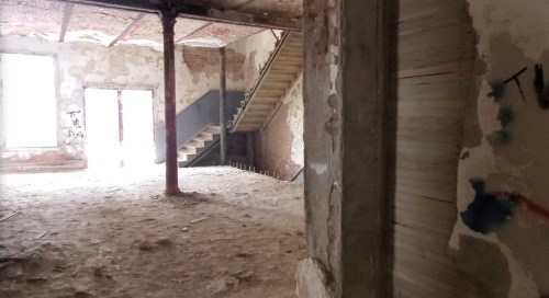 Abandoned interior of a building with exposed brick walls, a staircase, and debris on the floor. Natural light enters from a doorway.