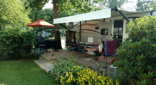 A cozy campsite with a trailer, outdoor seating, and vibrant flowers, surrounded by greenery and a shaded umbrella.