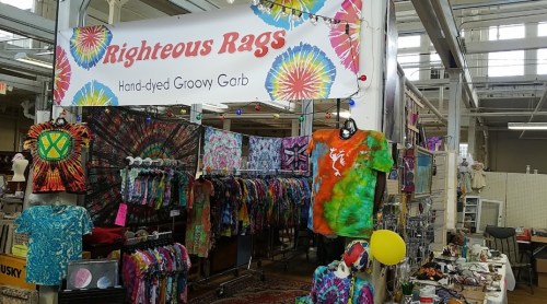 A colorful booth displaying hand-dyed tie-dye shirts and garments, with a sign reading "Righteous Rags."