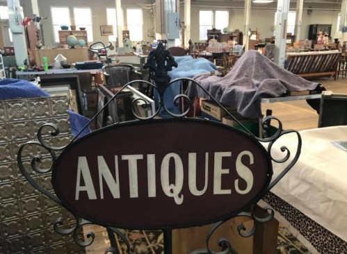A sign reading "Antiques" in a vintage shop filled with various furniture and items.