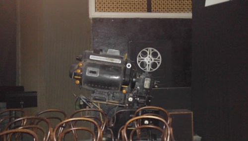 An old film projector positioned beside wooden chairs in a dimly lit room.