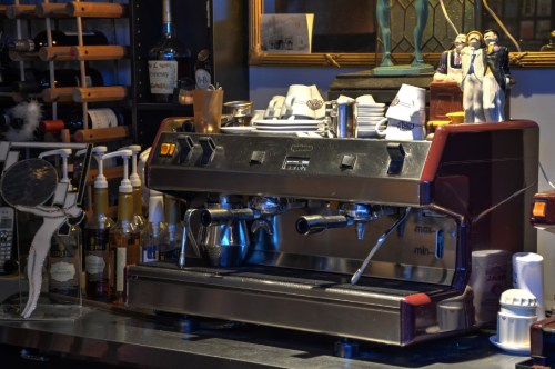 A vintage espresso machine on a bar counter, surrounded by bottles and coffee accessories.