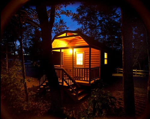 A cozy cabin illuminated at night, surrounded by trees, with warm light glowing from the windows.