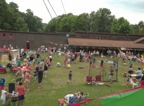 A lively outdoor gathering with people of all ages enjoying games, activities, and colorful decorations in a grassy area.