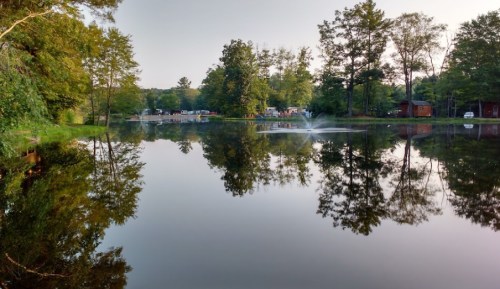 A serene lake reflecting trees and a fountain, with cabins visible in the background under a clear sky.