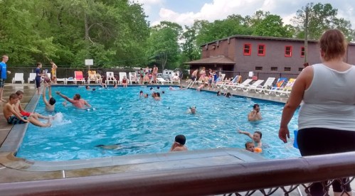 A busy outdoor pool scene with children and adults swimming and relaxing on lounge chairs under a sunny sky.