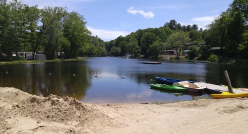 A serene lake surrounded by trees, with colorful kayaks on the shore and a clear blue sky above.