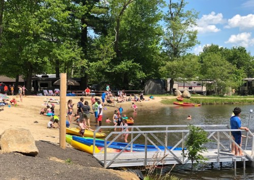 A sunny beach scene with people swimming, kayaking, and relaxing by a pond surrounded by trees and a sandy shore.