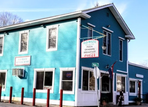 A blue two-story building with a sign for "Old Mystic" and a general store, featuring large windows and a red pole barrier.