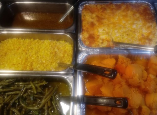 A variety of dishes in metal trays, including green beans, corn, sweet potatoes, and a cheesy casserole.