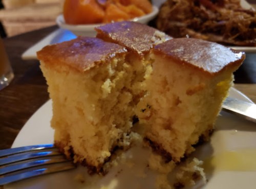 A close-up of a slice of moist cake on a plate, with a fork beside it and other food items in the background.