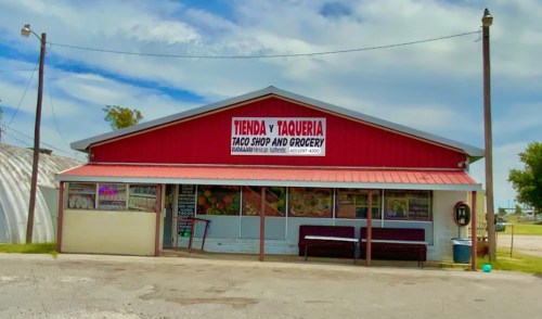 Red building with a sign reading "Tienda y Taqueria Taco Shop and Grocery," featuring a porch and outdoor seating.