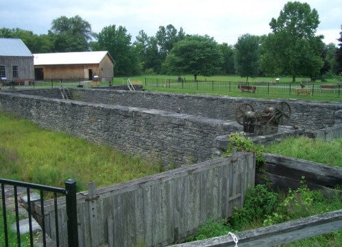 Stone ruins of a historic structure surrounded by greenery, with a wooden building in the background.