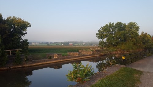 A serene landscape featuring a calm river, lush greenery, and a misty horizon under a clear sky.
