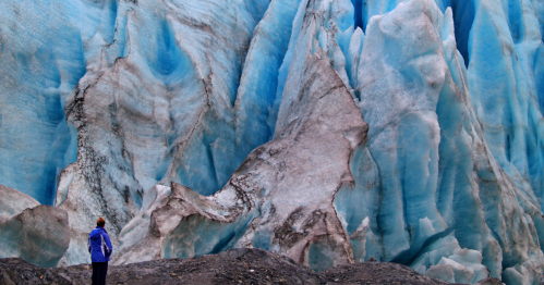 A person stands in front of a massive blue glacier, showcasing intricate ice formations and textures.