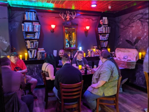 A cozy, dimly lit room with bookshelves, candles, and a group of people gathered around a table.