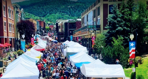A bustling street market with numerous tents, crowds of people, and mountains in the background on a sunny day.