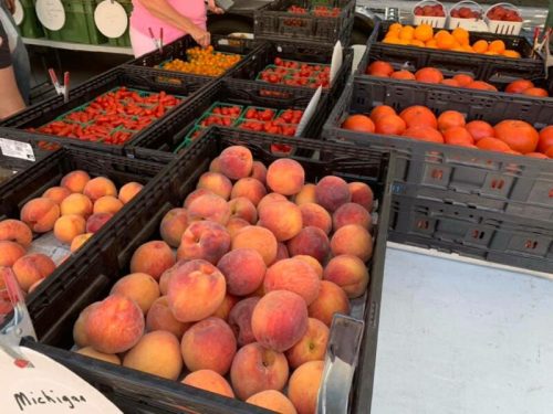 A market display featuring crates of peaches, tomatoes, and oranges, with a vendor in the background.