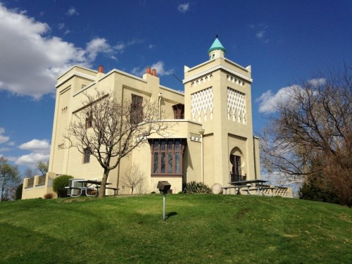 A unique, light-colored building with a tower and decorative elements, set on a grassy hill under a blue sky.