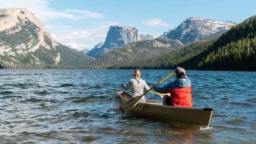 Two people paddle a canoe on a serene lake surrounded by mountains and lush forests under a clear blue sky.