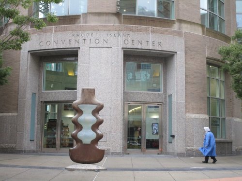 A person in a blue coat walks past the Rhode Island Convention Center, featuring a large abstract sculpture at the entrance.