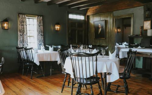 Cozy restaurant interior with wooden floors, white tablecloths, and vintage decor, featuring dark wooden chairs and soft lighting.