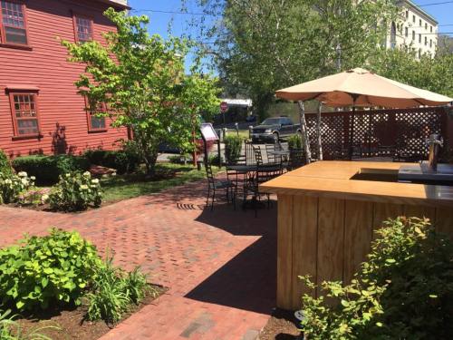A sunny outdoor patio with brick pathways, a wooden bar, tables, and greenery, surrounded by buildings and trees.