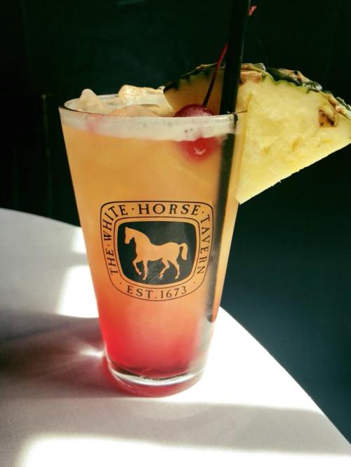 A colorful cocktail in a glass with a logo, garnished with a pineapple slice and a cherry, served on a table.