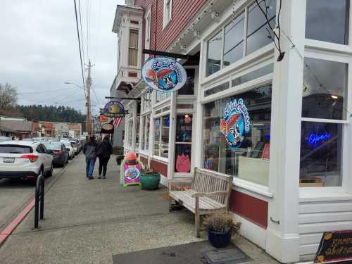 A colorful storefront with signs, featuring a bench outside and people walking on the sidewalk in a small town.