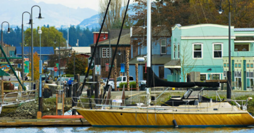 A yellow sailboat docked near colorful buildings and lampposts by the water, with mountains in the background.