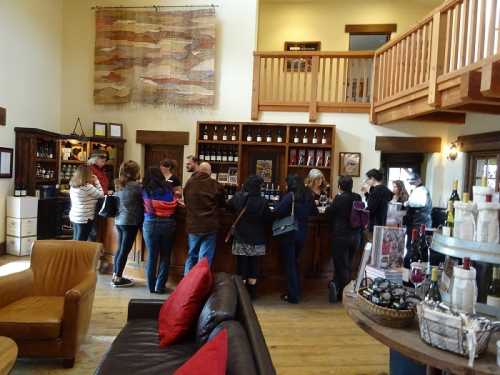 A busy wine tasting room with people at the bar and a cozy seating area in the foreground.