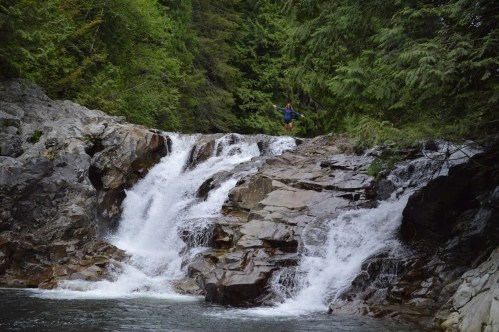 A person stands on rocky terrain beside a waterfall, surrounded by lush green trees and flowing water.