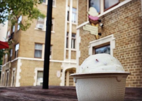 A close-up of a scoop of ice cream in a cup, with a brick building and sign in the background.