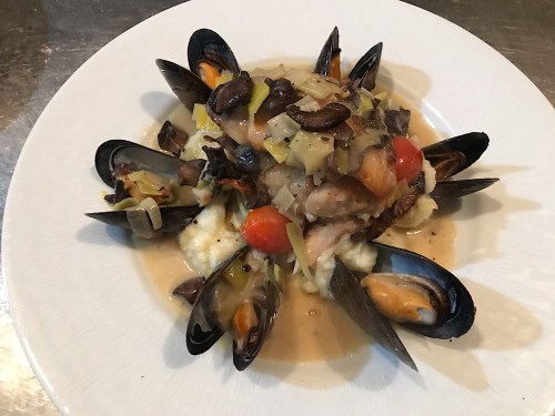 A plate of fish topped with mussels, vegetables, and sauce, served over creamy mashed potatoes.