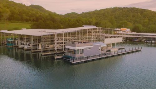 A marina with multiple boat docks on calm water, surrounded by green hills under a pastel sky.