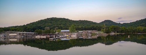 Scenic view of a calm lake with boats, docks, and lush green hills under a soft sunset sky.