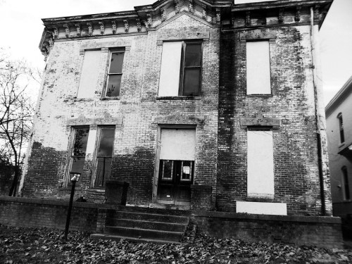 Abandoned brick house with boarded windows, showing signs of decay and overgrown foliage in black and white.