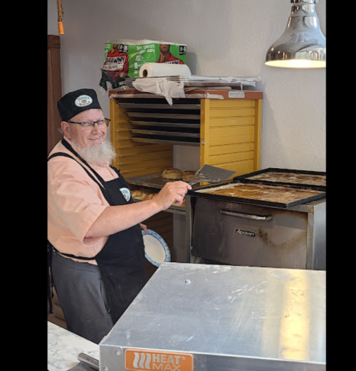 A smiling chef with a beard prepares food in a kitchen, wearing a black apron and a chef's hat.