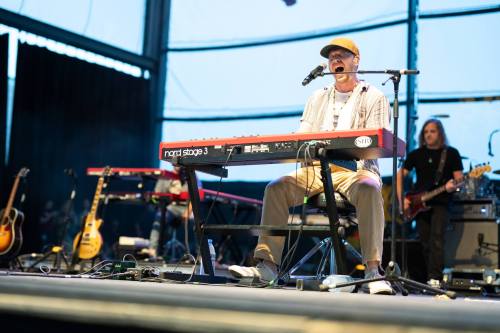 A musician performs on stage at a keyboard, with a band in the background and instruments visible.