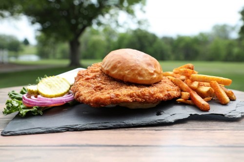 A crispy fried chicken sandwich with lettuce, pickles, and onion, served with golden fries on a slate platter.
