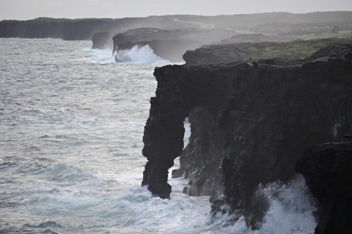 Rugged coastal cliffs with waves crashing against them, shrouded in mist and surrounded by a gray sky.