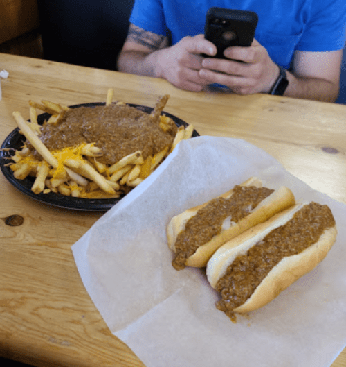 A plate of loaded fries topped with chili sits next to two chili dogs on a wooden table. A person is using a phone.